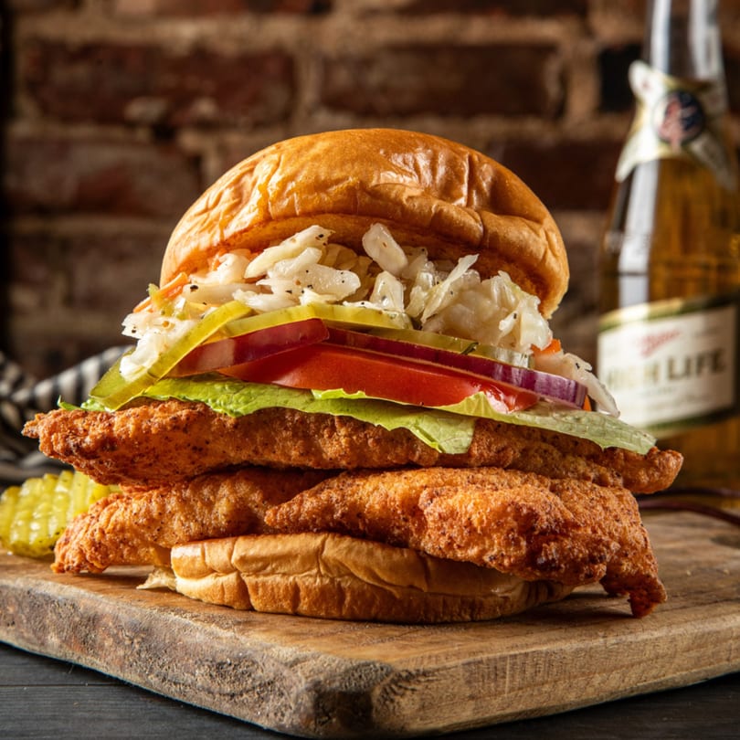 A large fried chicken sandwich with coleslaw, pickles, tomato, and lettuce on a bun.